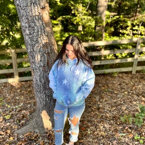 May include: A blue and white tie-dye hoodie with white stars printed on it. The hoodie has a drawstring hood and a kangaroo pocket. The person is wearing ripped jeans and white sneakers.