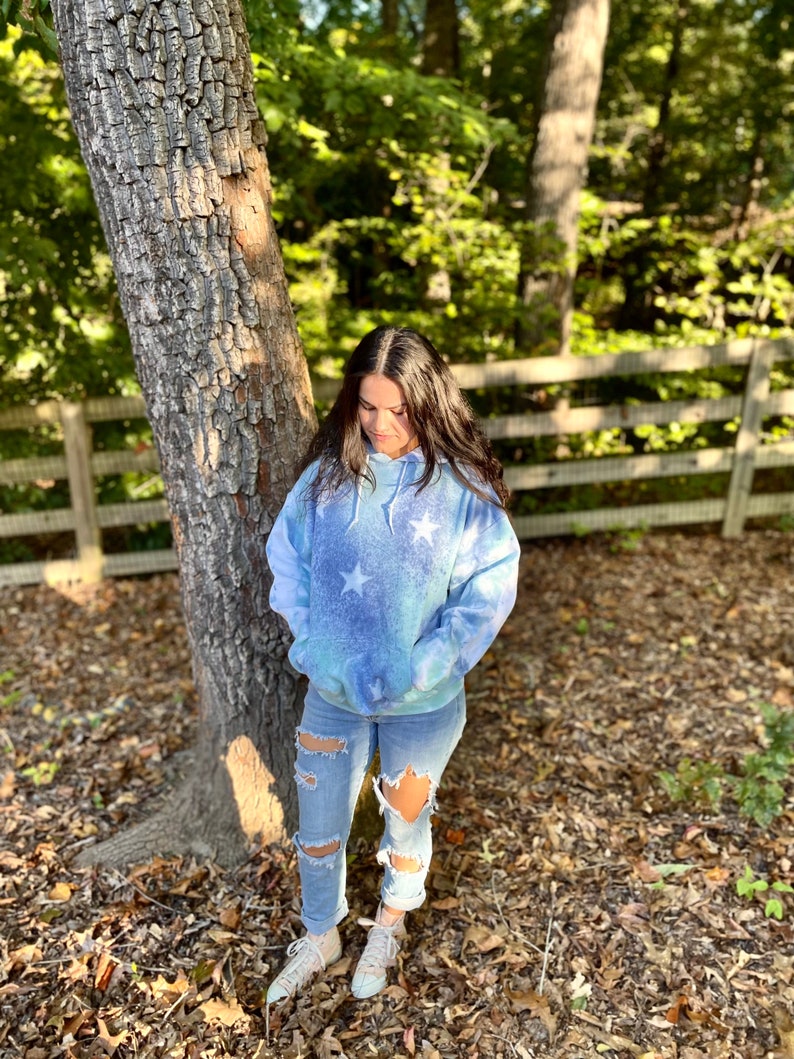 May include: A blue and white tie-dye hoodie with white stars printed on it. The hoodie has a drawstring hood and a kangaroo pocket. The person is wearing ripped jeans and white sneakers.