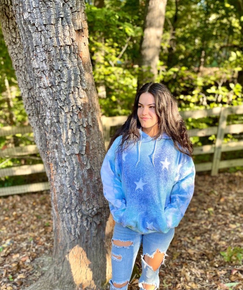 May include: A woman wearing a blue tie-dye hoodie with white stars. The hoodie has a drawstring hood and a kangaroo pocket. She is wearing blue jeans with distressed details.
