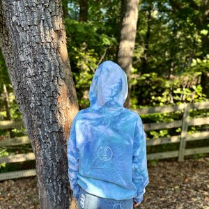 May include: A blue tie-dye hoodie with a white circle logo on the back. The hoodie is worn by a person standing in front of a tree and a wooden fence.