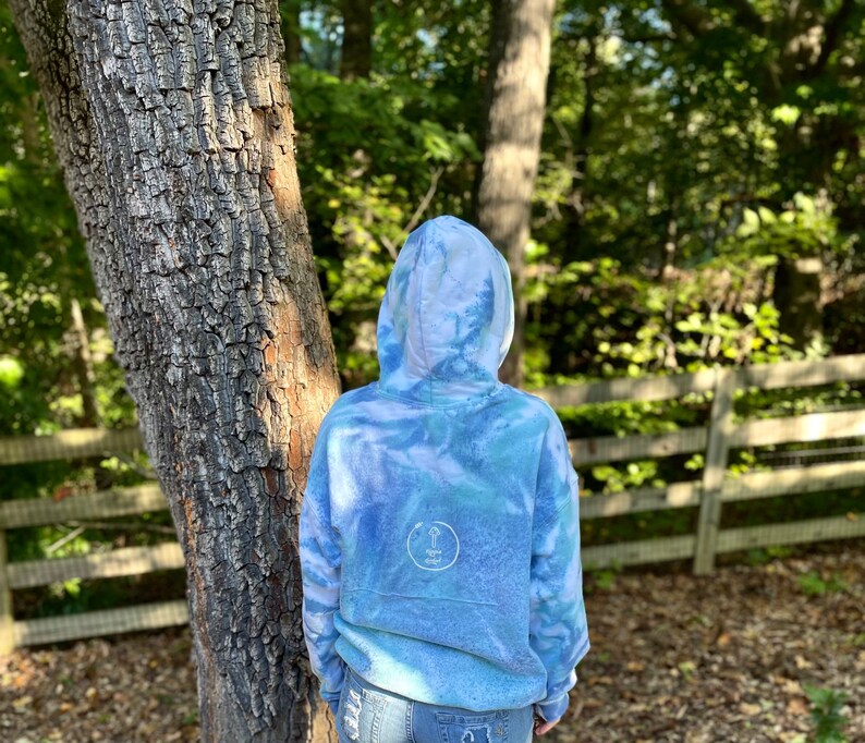 May include: A blue tie-dye hoodie with a white circle logo on the back. The hoodie is worn by a person standing in front of a tree and a wooden fence.