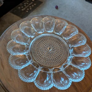 May include: A clear glass egg serving platter featuring twelve shell-shaped indentations arranged in a circular pattern around a central disc with a textured hobnail design. The edges of the platter are scalloped, and the glass is transparent.