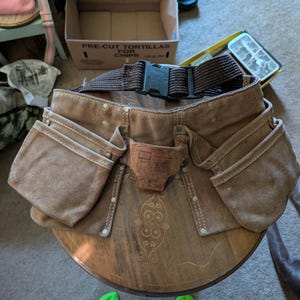 May include: A brown canvas tool belt featuring multiple storage pouches, a central leather hammer loop, and a dark brown adjustable nylon webbing strap with a black plastic side-release buckle. The belt is constructed with reinforced metal rivets.