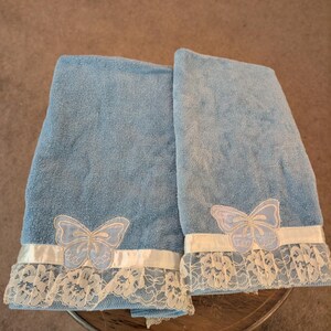 May include: Two folded light blue terry cloth towels, each featuring a white satin ribbon trim along the bottom edge. A white embroidered butterfly appliqué is centered on the ribbon, and a delicate white floral lace border finishes the hem.