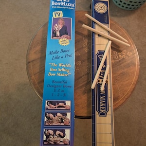 Op de afbeelding: Een Deluxe E-Z BowMaker-set in een blauwe kartonnen doos met producttekst en afbeeldingen, naast een lichtgekleurde houten basis met een bedrukte liniaal en diverse verwijderbare houten deuvels voor het maken van strikken, inclusief spoelhouder.