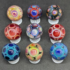 May include: Nine spherical glass marbles arranged in a three-by-three grid. Each marble features a distinct multi-colored pattern of concentric circles and swirling glass layers in shades of blue, red, yellow, orange, black, and iridescent white.