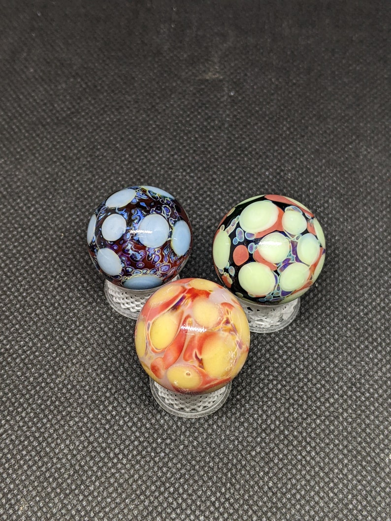 Puede incluir: Tres canicas de vidrio con patrones abstractos coloridos. Las canicas son rojas, amarillas y azules con acentos blancos. Las canicas est&aacute;n sobre un fondo gris oscuro.