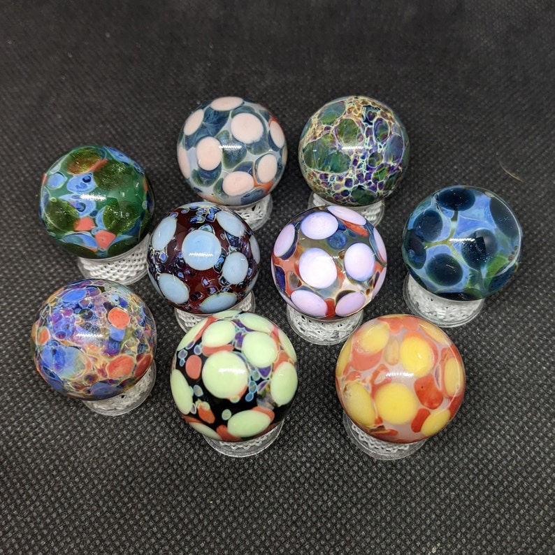 Puede incluir: Nueve canicas de vidrio de colores con intrincados patrones, cada canica se muestra en un peque&ntilde;o soporte blanco.