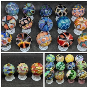 May include: A collection of spherical glass marbles featuring intricate, multi-colored patterns with swirling designs. Each marble is displayed on a small, white, circular, lattice-patterned stand, showcasing various color combinations including blue, orange, green, and black.