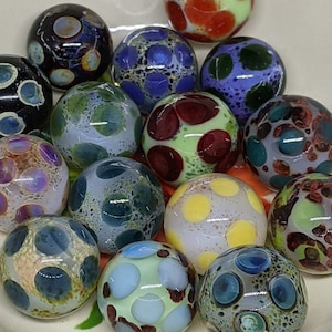 Puede incluir: Una colecci&oacute;n de 14 canicas de vidrio de colores con varios patrones y colores, incluyendo azul, verde, rojo, blanco y negro.