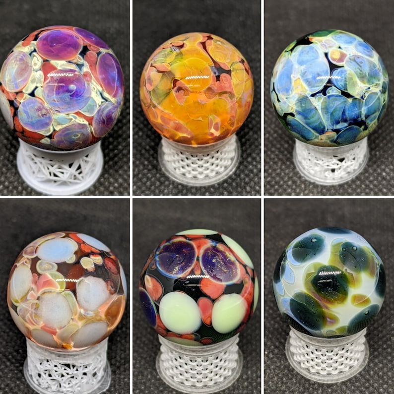 Puede incluir: Seis canicas de vidrio con patrones abstractos coloridos. Cada canica se muestra en un soporte blanco.