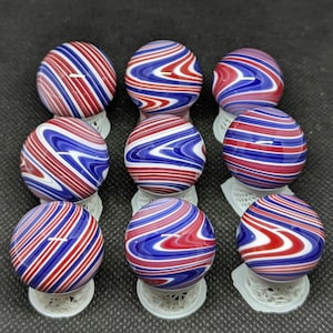 May include: Nine glass marbles with red, white, and blue swirling patterns. Each marble is supported by a small, white, lace-like stand. The marbles have a glossy finish and are arranged in a grid.