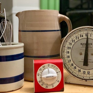 May include: A vintage kitchen scale with a white face and black numbers, a red kitchen timer, a white ceramic pitcher with blue stripes, and a white ceramic crock with blue stripes filled with kitchen utensils.