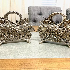 May include: Two silver napkin holders shaped like grapevines with grapes. The holders are on a wooden table.