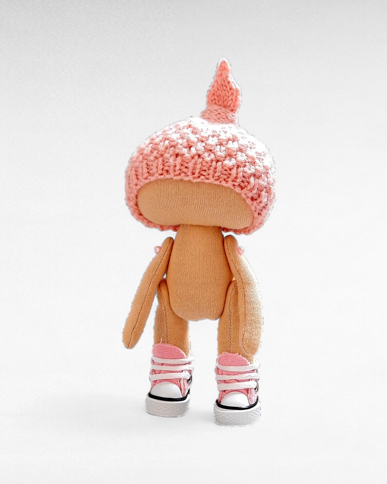Op de afbeelding: Een handgemaakte pop met een roze gebreide muts en roze sneakers. De pop heeft een beige stoffen lichaam en staat op een witte achtergrond.
