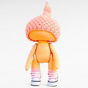 Op de afbeelding: Een handgemaakte pop met een roze gebreide muts, een oranje stoffen lichaam en roze sneakers.