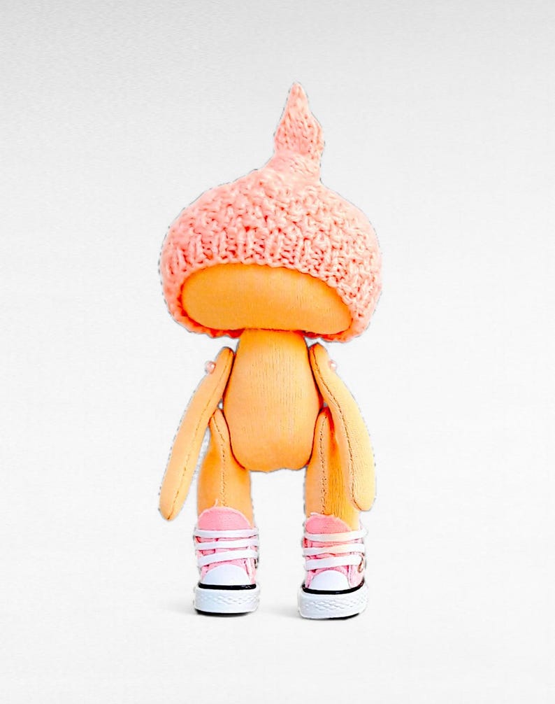 Op de afbeelding: Een handgemaakte pop met een roze gebreide muts, een oranje stoffen lichaam en roze sneakers.