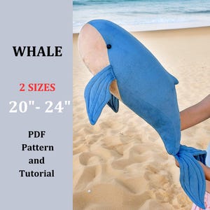 May include: A blue and tan plush whale toy, with the word "WHALE" in black. The text "2 SIZES 50-61 cm" is visible, along with "PDF Pattern and Tutorial". The whale is held up against a beach background.