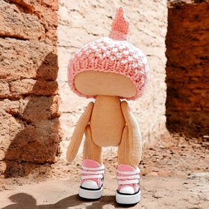 Op de afbeelding: Een handgemaakte pop met een roze gebreide muts en roze en witte sneakers. De pop staat op een bruin oppervlak.