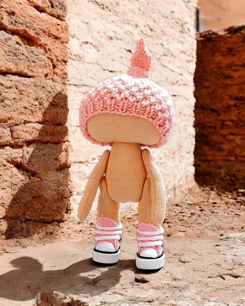 Op de afbeelding: Een handgemaakte pop met een roze gebreide muts en roze en witte sneakers. De pop staat op een bruin oppervlak.