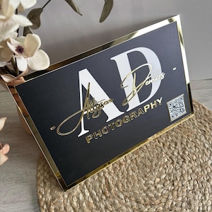 May include: Black and gold sign with the letters "AD" in white, the text "Alyson Demers" and "PHOTOGRAPHY" in white, and a QR code.