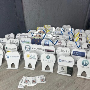 May include: A collection of white tooth-shaped promotional stands with various dental practice logos and names. The stands are arranged on a wooden surface with a few business cards and small stickers with QR codes.