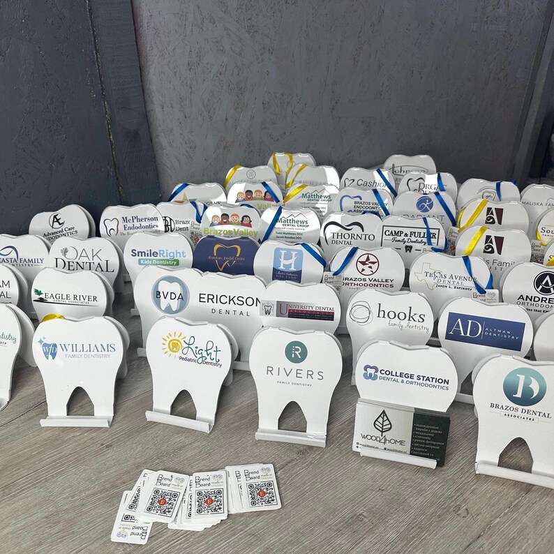 May include: A collection of white tooth-shaped promotional stands with various dental practice logos and names. The stands are arranged on a wooden surface with a few business cards and small stickers with QR codes.