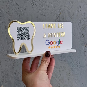 Peut inclure: Panneau blanc pour les avis dentaires avec une forme de dent dorée et un code QR. Le panneau indique "LAISSEZ-NOUS UN AVIS Google" avec cinq étoiles dorées. Le panneau est soutenu par une étagère blanche.