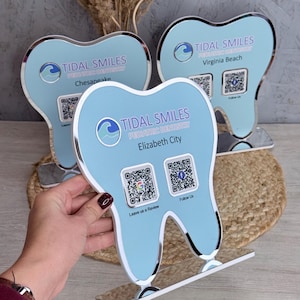 May include: Three light blue tooth-shaped signs with silver trim. Each sign has the text "TIDAL SMILES PEDIATRIC DENTISTRY" and a city name: Chesapeake, Elizabeth City, and Virginia Beach. Each sign has two QR codes.