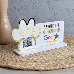 May include: A white and gold paw print-shaped sign with the text "LEAVE US A REVIEW Google" and a QR code. The sign is on a white base and sits on a woven mat. The Google logo is in blue and gold.