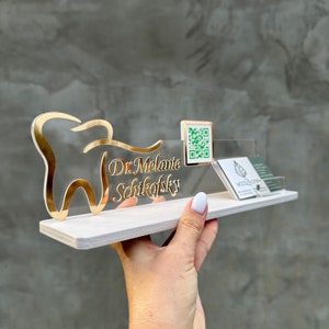 Könnte beinhalten: Ein weißes Acryl-Schreibtisch-Namensschild mit einem goldenen Zahndesign und dem Text "Dr. Melanie Schickofsky". Ein kleines Quadrat mit einem QR-Code und ein Visitenkartenhalter sind ebenfalls enthalten.