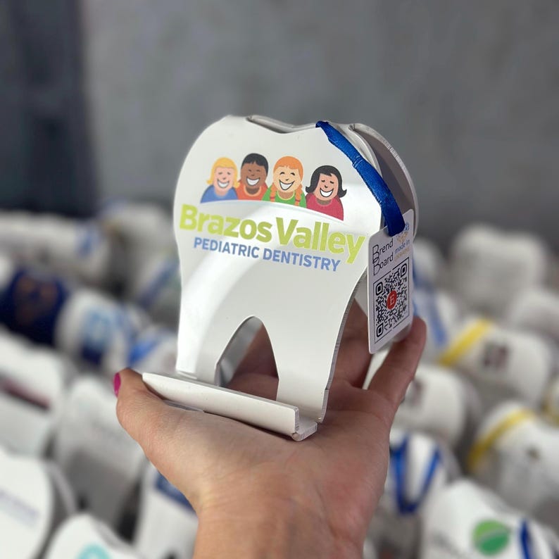 May include: White plastic tooth-shaped stand with a blue ribbon and a QR code. The stand has a graphic of four children and the text "Brazos Valley Pediatric Dentistry".
