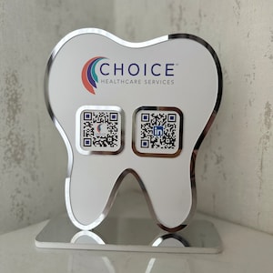 May include: A white tooth-shaped sign with a silver border. The sign has the text "CHOICE" in blue and "HEALTHCARE SERVICES" in black. There are two QR codes on the sign, one with the word "CHOICE" and the other with the LinkedIn logo.