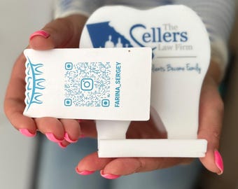 May include: White business card with a blue and white QR code and the text "FARINA_SERGEY" on a white tooth-shaped stand. The stand has the text "The Sellers Law Firm" and "Hearts Become Family" on it.