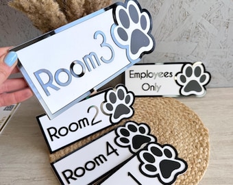 May include: A collection of rectangular white signs with black borders and a black paw print icon. The signs feature black text reading Room 1, Room 2, Room 3, Room 4, and Employees Only. Each sign has a reflective finish and a layered design.