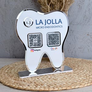 Könnte beinhalten: Ein weißes, zahnförmiges Acrylschild mit verspiegeltem Rand und Sockel. Es zeigt den Text LA JOLLA MICRO ENDODONTICS, ein kreisförmiges LJ-Logo sowie zwei QR-Codes, die mit den Symbolen für Instagram und Google Reviews versehen sind.