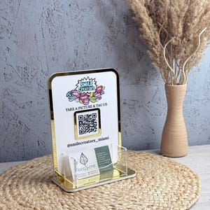 Peut inclure: Un porte-cartes de visite en acrylique blanc et doré avec le texte "SMILE CREATORS" et un code QR. Le support a une bordure dorée et une poche en acrylique transparente pour les cartes. Le texte "TAKE A PICTURE & TAG US" est également visible.