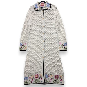 Könnte beinhalten: Cremefarbener Häkel-Duster mit floralen Stickereien an Kragen, Manschetten und Saum. Die langärmelige Strickjacke hat eine Knopfleiste und schwarze Paspeln. Die Blumenstickerei umfasst rosa, blaue und gelbe Blumen.