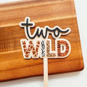 Peut inclure: Topper de g&acirc;teau "Two Wild" avec une police d'&eacute;criture cursive noire pour le mot "two" et le mot "WILD" en lettres imprim&eacute;es d'animaux brunes, orange et noires.