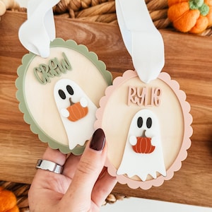 May include: Two Halloween-themed wooden ornaments. One is light green with the word "CREW" and a ghost holding a pumpkin. The other is light pink with the name "RYLIE" and a ghost holding a pumpkin. Both have white ribbons.