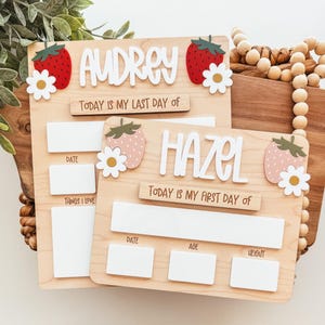 May include: Two wooden milestone boards with the names "Audrey" and "Hazel" in white letters. Each board features strawberry and daisy accents. Text includes "Today is my last day of" and "Today is my first day of".