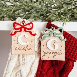 Peut inclure: Deux étiquettes de bas de Noël en bois avec un motif de nœud. L'une a un nœud rouge et le nom "Cecelia", l'autre a un nœud vert et le nom "Georgia". Les deux étiquettes ont une initiale blanche et des étoiles.