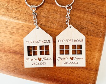 May include: Two wooden house-shaped keychains with silver key rings. Each keychain has the text "Our First Home" and the names "Hussein" and "Touma" with a heart symbol between them. The date "28.02.2023" is also engraved on each keychain.