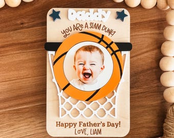 May include: Wooden Father's Day gift featuring a basketball hoop design with a photo of a baby. The text reads "Daddy, you are a slam dunk!" and "Happy Father's Day! Love, Liam."