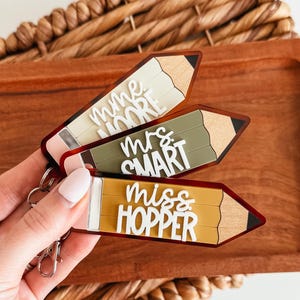 May include: Three pencil-shaped keyrings with teacher names: "Mme Moore", "Mrs. Smart", and "Miss Hopper". The keyrings are in beige, olive green, and gold, with white lettering and a pencil tip design. They are attached to a silver keyring.