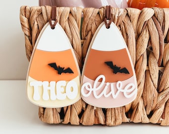 May include: Two candy corn shaped decorations with the names Theo and Olive written on them. Each decoration has a black bat silhouette on the orange section.