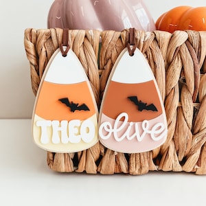 May include: Two candy corn shaped ornaments with the names Theo and Olive written on them. Each ornament has a black bat silhouette on the orange section.