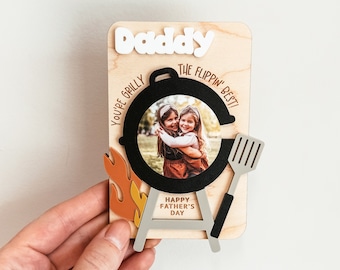 May include: A wooden Father's Day card with a photo frame shaped like a grill. The card says "Daddy, You're Grilly the Flippin' Best!" and "Happy Father's Day".