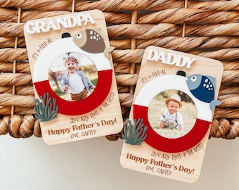 May include: Two wooden Father's Day cards with a fishing theme. Each card has a photo frame shaped like a life preserver with a fish on top. The text on the cards reads "It's O-Fish-Al" and "You Are Reel-Y The Best! Happy Father's Day! Love, Carter".