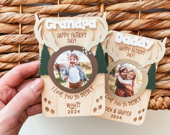 May include: Two wooden photo frames with a deer antler design. The frames have a photo of a child in each. The text on the frames reads "Grandpa Happy Father's Day! I Love You So Deerly Wyatt 2024" and "Daddy Happy Father's Day! I Love You So Deerly Den & Harper 2024".